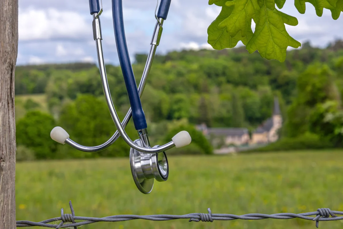 Stethoscope hanging on a barbed-wire fence at the edge of a rural field, a distant village behind it, evoking the healthcare provider shortage in rural areas