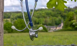 Stethoscope hanging on a barbed-wire fence at the edge of a rural field, a distant village behind it, evoking the healthcare provider shortage in rural areas