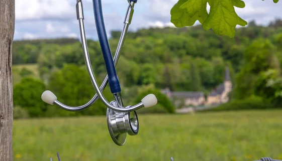 Stethoscope hanging on a barbed-wire fence at the edge of a rural field, a distant village behind it, evoking the healthcare provider shortage in rural areas