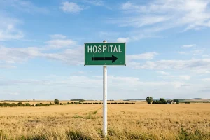 A green HOSPITAL road sign with a directional arrow stands in an open field of farmland, the kind of rural community where provider vacancies and a long time-to-hire hit hardest